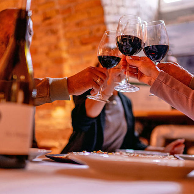 Drei Personen stoßen in gemütlicher Atmosphäre mit Rotweingläsern an, im Hintergrund warmes Licht und ein gedeckter Tisch.