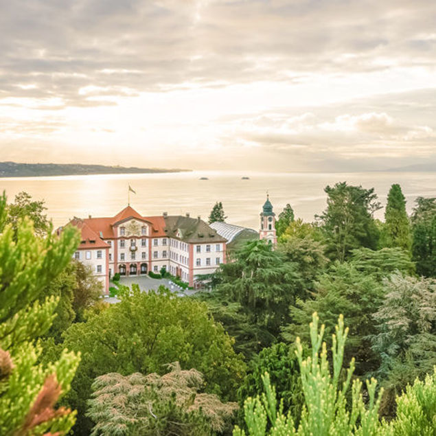 Blick über eine grüne Parklandschaft auf ein historisches Gebäude am Ufer eines Sees, im Hintergrund ruhiges Wasser und bewölkter Himmel im Abendlicht.