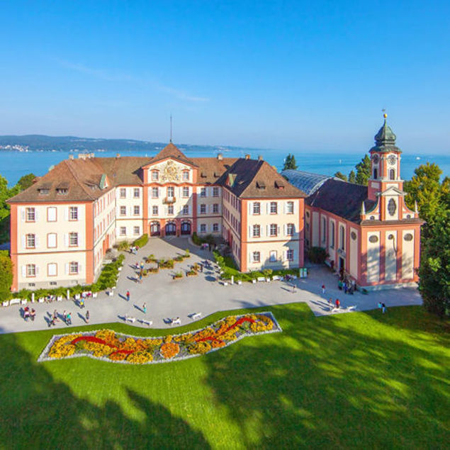 Luftaufnahme von Schloss Mainau mit Schlosskirche, großem Innenhof und farbenfrohem Blumenbeet, im Hintergrund der Bodensee