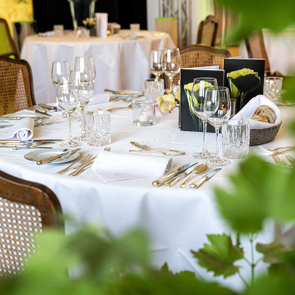 Ein gedeckter Tisch mit edler weißer Tischkette, Silberbesteck und verschiedene Weingläser. Im Vordergrund bilden Weinblätter einen Rahmen um die Szenerie