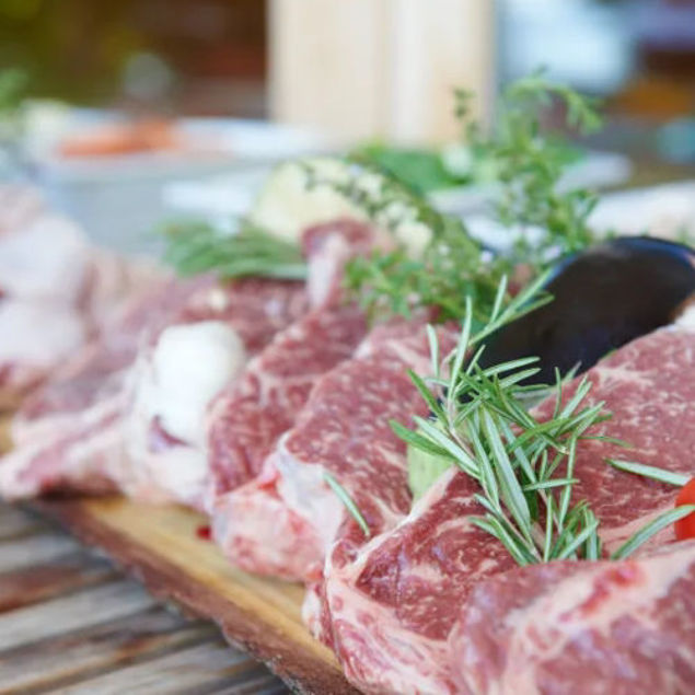 Mehrere rohe, marmorierte Rindersteaks liegen auf einem Holzbrett, garniert mit frischen Kräutern wie Rosmarin; das Bild ist nah aufgenommen und der Hintergrund unscharf.