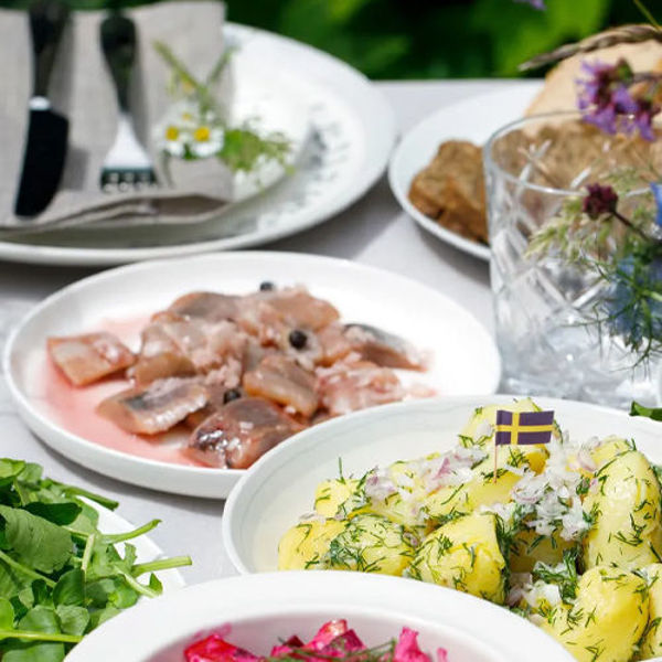 Gedeckter Tisch mit verschiedenen Speisen, darunter Kartoffelsalat mit Dill, eingelegter Fisch und frische Kräuter, dekoriert mit Blumen