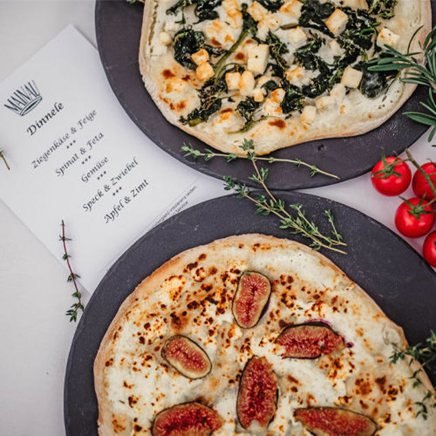 Zwei Pizzen auf dunklen Tellern, eine mit Tomaten und Käse, die andere mit grünem Belag, daneben Kräuter und Kirschtomaten