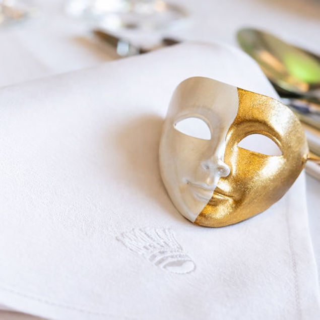 Eine goldene, dekorative Theatermaske liegt auf einer weißen Stoffserviette