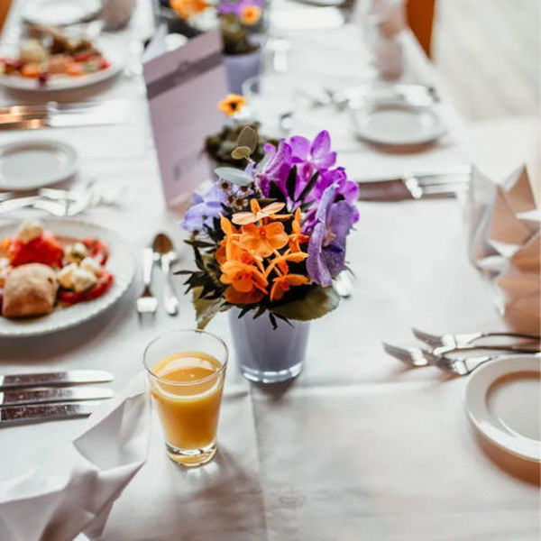 Ein festlich gedeckter Tisch strahlt eine elegante, zugleich einladende Atmosphäre aus. Auf der weißen Tischdecke sind Teller, glänzendes Besteck und sorgfältig gefaltete Servietten harmonisch arrangiert. Im Mittelpunkt steht ein kleines, rundes Blumenarrangement in einer zarten Vase: Leuchtendes Orange, kräftiges Violett und sanftes Lila setzen farbenfrohe Akzente und bringen Lebendigkeit in das helle Gesamtbild.